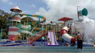 Keluarga sedang bermain ceria di taman hiburan air dengan seluncuran dan kolam renang yang ramai.