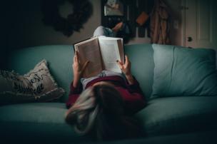 Seseorang sedang membaca buku di sofa nyaman di sudut cafe yang penuh buku.
