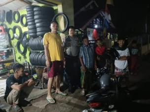 Gambar tempat berkumpul komunitas motor yang ramai, dengan berbagai jenis motor terparkir, orang-orang bercakap-cakap, dan area bengkel kecil di latar belakang.
