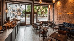 Interior coffee shop yang estetik dengan pencahayaan alami yang masuk melalui jendela besar, terlihat beberapa spot duduk yang nyaman.